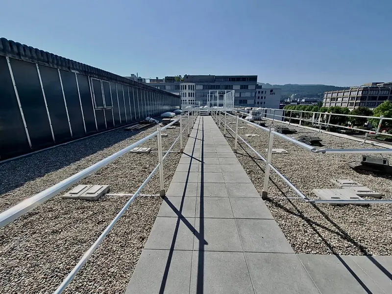 Flachdach mit Kiesschüttung: Plattenweg in der Bildmitte, beidseitig Aluminium-Geländer auf Betonfüssen, im Hintergrund Bürogebäude.