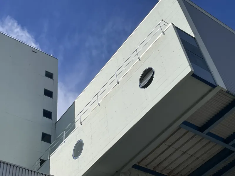 Blick nach oben auf einen auskragenden Sichtbetonbau mit zwei runden Fenstern und Geländer auf der Dachkante; links ein hoher Gebäudeteil, dazu blauer Himmel.