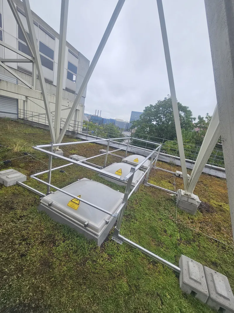 Begrüntes Flachdach mit drei Lichtkuppeln hintereinander, jede mit Aluminium-Geländerrahmen; vorn weisse V-förmige Stahlstreben.