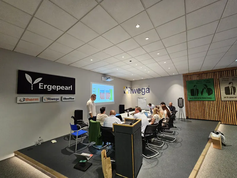 Schulungsraum mit den Logos Ergepearl, 3therm, Riwega und RoofRox an grauer Wand; Teilnehmende an einem langen Tisch verfolgen eine Beamer-Präsentation.
